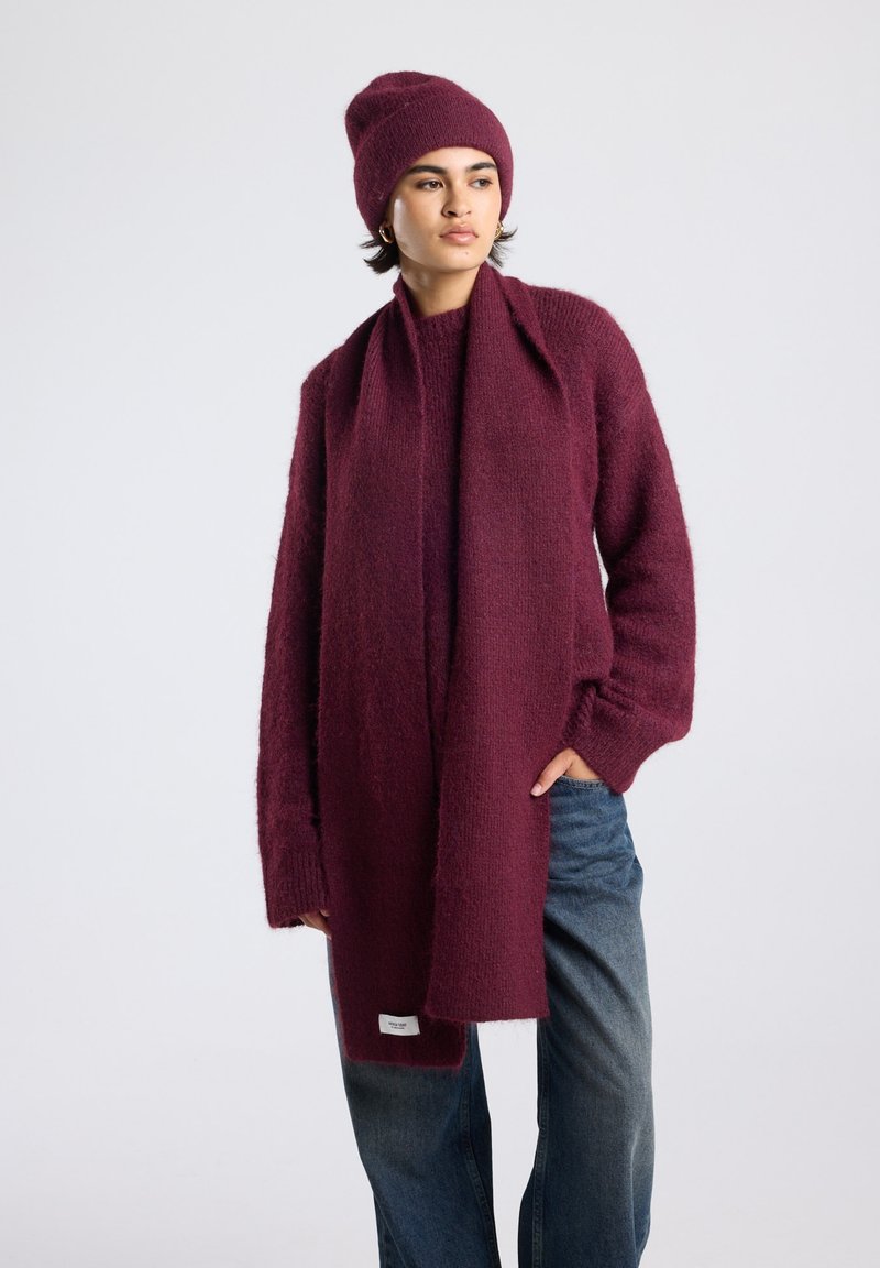 Burgundy knitted cardigan and scarf made from soft, textured material, paired with a matching beanie. Casual fit, relaxed sleeves, denim jeans.