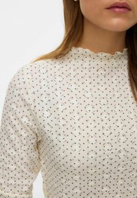 Beige blouse met een hoge, gegolfde halslijn met een gestructureerd patroon van kleine gaatjes en contrasterende stippen in bruin en crème.