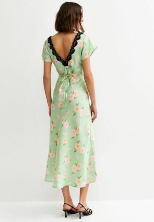 Vrouw draagt een lichtgroene midi-jurk met bloemenprint, korte mouwen, zwarte kanten V-rugdetail, taille met strik en zwarte sandalen met hak.