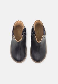 Para czarnych skórzanych butów wsuwanych z brązowym wnętrzem i falistymi krawędziami, widziana z góry na białym tle.