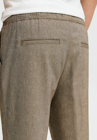 Close-up of beige textured trousers with two back welt pockets and elastic waistband, worn with a white casual shirt.