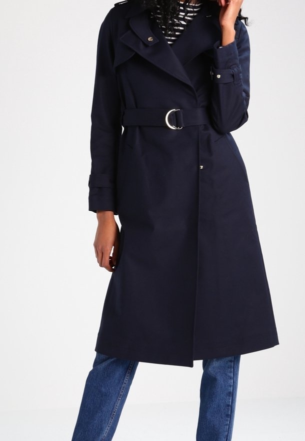 Kvinna iklädd en marinblå bälte trench coat över en randig topp och blå jeans, stående med ena handen nära ansiktet och den andra avslappnad.