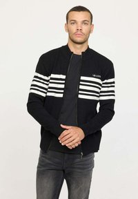 Sweatshirt zippé noir à manches longues, avec des accents rayés blancs sur la poitrine. Fabriqué en tissu texturé avec un design ajusté.