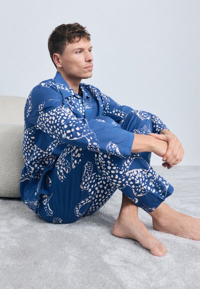 Blå pyjamas sæt med et leopardmønster i hvid. Har lange ærmer og bukser, lavet af et glat stof. Siddende på et gråt gulv.