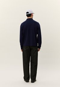 Mann sett bakfra iført hvit caps, marineblå langermet skjorte, svarte vide bukser og svarte sko på en ensfarget lys bakgrunn.