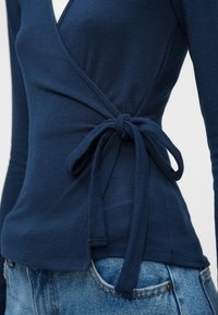 Tummansininen kääntötoppi pitkillä hihoilla ja sivusiteellä. Pehmeä rakenne, istuva siluetti ja syvä V-pääntie. Yhdistetty vaaleisiin farkkuihin.