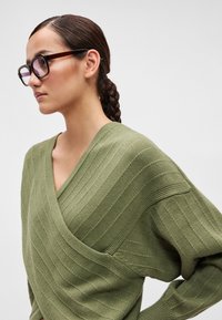 Ελιάς πράσινο πλεκτό παλτό με ραφη περιθώριο και βαθύ V-λαιμόκοψη. Χοντρές, χαλαρές μανσέτες συμπληρώνουν τον ριχτό σχεδιασμό.