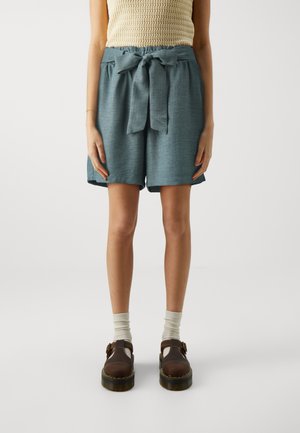 Teal linned shorts med en bred talje, bundet i en sløjfe, der har en afslappet pasform og sidelommer, parret med brune sko med spænder og lyse sokker.