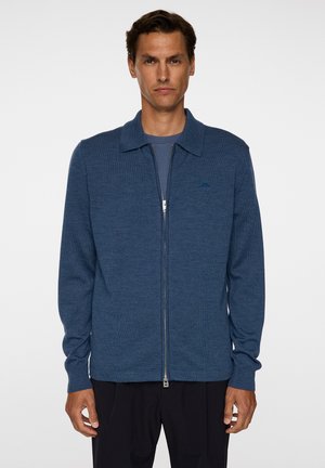 Blå lynlås cardigan lavet af blødt strikket stof, med flad krave, lange ærmer og en diskret logo detalje på brystet.