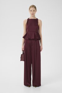 Žena v bezrukávovom burgundskom saténovom topi s peplum detailom a zodpovedajúcich širokých nohaviciach, drží malú textúrovanú kabelku.