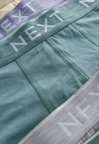 Sous-vêtements en coton vert, lavande et crème. Texture douce avec des ceintures élastiques contrastées portant le logo "NEXT" en lettres grasses.