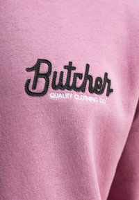 Helle rosa Sweatshirt mit besticktem schwarzem Text "Butcher" und "QUALITY CLOTHING CO". Glattes Material mit geripptem Struktur.