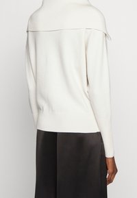 Crèmekleurige gebreide trui met een hoge halslijn en geribbelde afwerking. De stof ziet er zacht uit met een losse pasvorm en een lage rug.