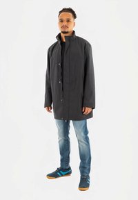 Vīrietis melnā ar rāvējslēdzēju jaku, zilos džinsos un zilos sporta apavos ar gaiši zilām svītrām, stāvot pret baltu fona.