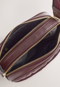 Burgunderfarvet lædertaske i crossbody-stil med tekstureret overflade, guldlynlåse og en sort indvendig foring med flere rum.