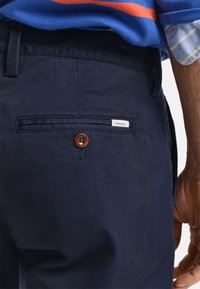 Pantalones de algodón azul marino con un bolsillo trasero que presenta un botón marrón y una etiqueta blanca de GANT cosida encima. Tejido texturizado, diseño a medida.