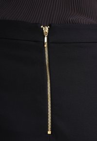 Černá sukně s vertikálním zlatým zipem v přední části, vyrobená z hladkého materiálu s elegantním a přiléhavým střihem.