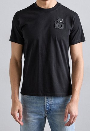 Mann trägt ein schwarzes T-Shirt mit einer kleinen schwarzen Schlangengrafik auf der linken Brust, kombiniert mit hellblauen Jeans.