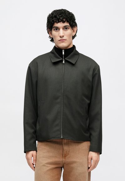 Donkergroene vest met rits, een kraag en lange mouwen, met een gladde textuur en zijzakken, gedragen over lichtbruine broek.