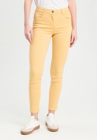 Jean skinny en denim jaune à taille haute, avec un design à cinq poches et une fermeture à bouton, associés à des baskets blanches avec des accents beiges.