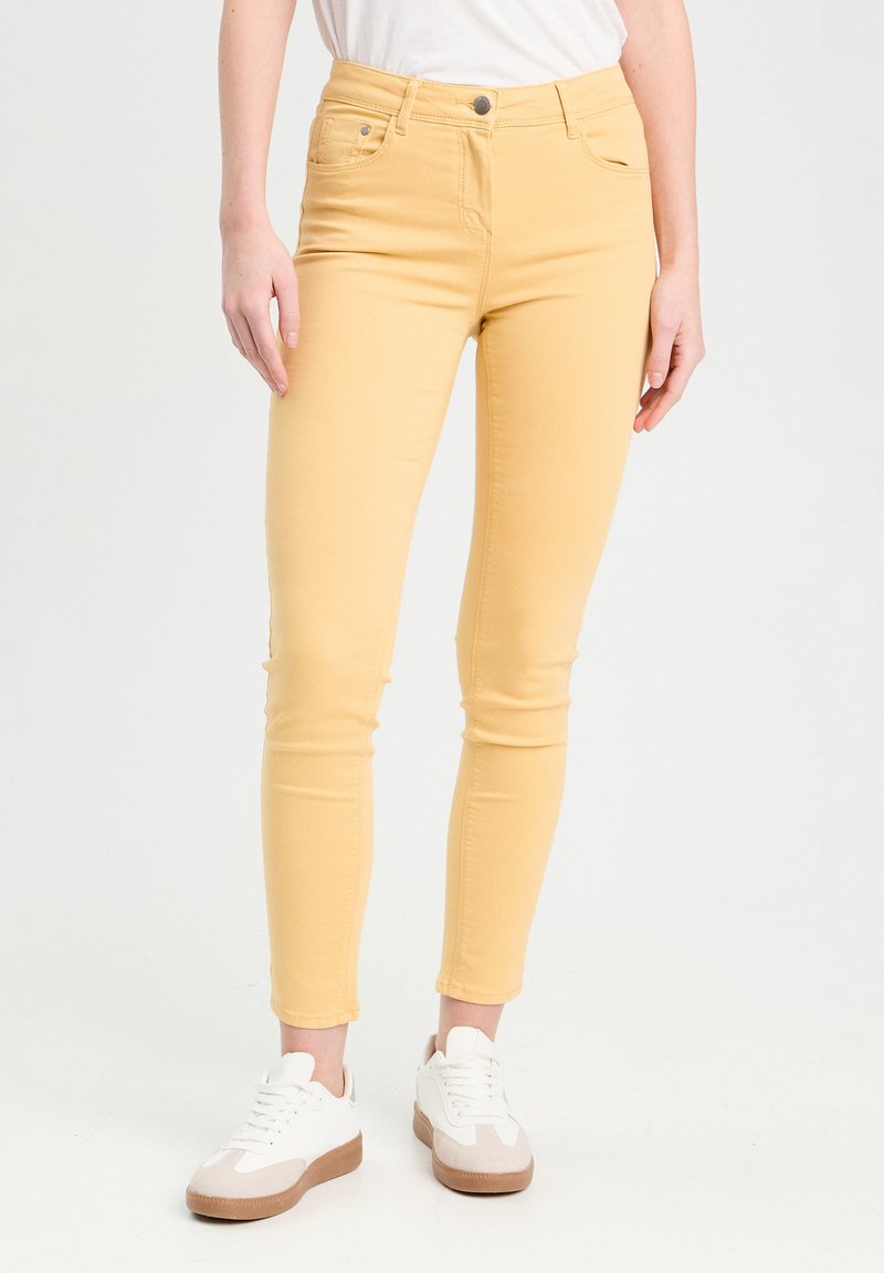 Jean skinny en denim jaune à taille haute, avec un design à cinq poches et une fermeture à bouton, associés à des baskets blanches avec des accents beiges.