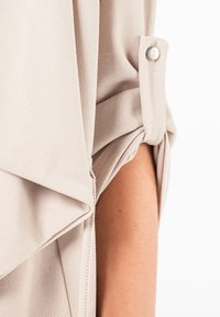 Blouse beige avec un design drapé, manches roulées maintenues par un système de bouton et une texture lisse.
