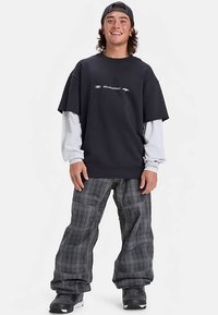 Mladý muž se usmívá, má na sobě šusťákovou čepici obrácenou vzad, černé a šedé vrstvené tričko s dlouhým rukávem, volné kostkované kalhoty a černé sněhové boty, stojí na bílém pozadí.