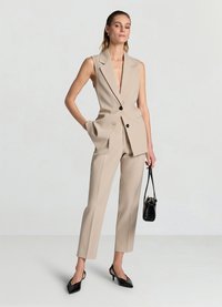 Ensemble veste sur mesure beige avec col en V profond, poches latérales et boutons noirs. Accessoirisé avec des talons aiguilles noirs et un petit sac à main noir.