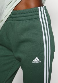 Gröna joggers i bomullsblandning med ribbad midja, prydda med tre vita ränder på sidorna och en broderad Adidas-logotyp.