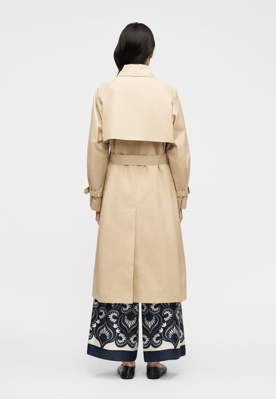 Γυναίκα με μακριά σκούρα μαλλιά, φορώντας μπεζ καμπαρντίνα πάνω από παντελόνι φαρδύ με σχέδια και μαύρα παπούτσια, στέκεται γυρισμένη με την πλάτη σε μονόχρωμο φόντο.