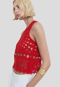 Red crochet halter top featuring openwork patterns and scalloped edges. Paired with white trousers and a curved gold bracelet.