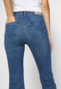 Siniset farkut, joissa on leveä lahje ja takataskut, joissa on kaareva sauma. Valmistettu teksturoidusta puuvillakankaasta, keski-korkean vyötärön istuvuus.