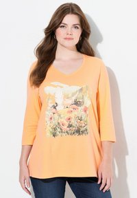 Pfirsichfarbene V-Ausschnitt Langarmbluse mit floralem Design, das Mohnblumen und eine Vogelillustration auf der Vorderseite zeigt. Weicher, leichter Stoff.