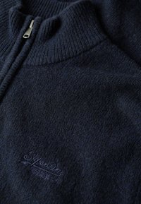 Maglione navy in morbido maglione con colletto alto e chiusura con zip. Caratterizzato da una sottile trama e logo ricamato "SuperDry" sul lato sinistro anteriore.