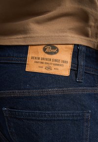 Vaqueros de mezclilla de color azul oscuro con una etiqueta de cuero marrón que muestra "Petrol" y "Denim Driven Since 1989". Presenta costuras visibles y un bolsillo trasero.