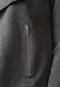 Tummanharmaa mokkainen takki, jossa pystysuora hopeanvärinen vetoketjutasku rinnassa ja näkyvät ompelusaumat.