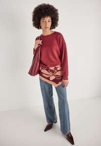 Felpa rossa, gonna mini con fantasia beige e bordeaux, jeans a gamba larga azzurri chiari e scarpe bordeaux a punta. Tiene una borsa rossa.