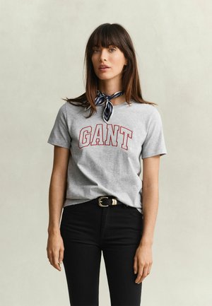 Femme aux cheveux bruns portant un t-shirt gris GANT, un pantalon noir avec ceinture et un foulard rayé autour du cou, debout devant un fond clair uni.