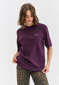 Fialové bavlněné tričko s krátkým rukávem a kulatým výstřihem. Na hrudi je menší bílý grafický motiv řas. Hladký materiál.
