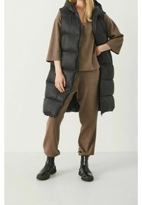 Persoon in een bruine gebreide set met brede mouwen, een zwarte lange gewatteerde vest en zwarte enkellaarsjes staande op een witte vloer.