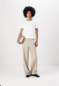 T-shirt bianco a maniche corte con un piccolo logo, abbinato a pantaloni beige a gamba larga. Il modello tiene una borsa grigia chiaro.