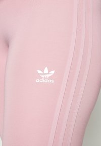 Rosa leggings i mjukt tyg med en vit Adidas-logotyp och tre vita ränder längs sidan.