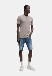 Polo beige con maniche corte, abbinato a pantaloni shorts di denim azzurro chiaro. Modello che indossa sneakers bianche e calzini bianchi alla caviglia.