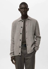 Szary prążkowany kardigan z kołnierzykiem, zapinany na guziki z przodu, z długimi rękawami. Noszony na ciemnozielonej koszuli, w połączeniu z dopasowanymi spodniami.