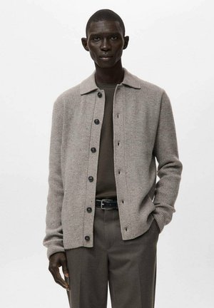 Cárdigan gris acanalado con diseño de cuello, botones al frente y mangas largas. Llevado sobre una camisa verde oscuro, combinado con pantalones de corte ajustado.