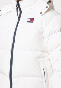 Hvid dunjakke med lynlås foran og marineblå detaljer. Features inkluderer hætte, sidelommer og en logopatch på brystet.