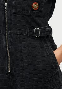 Κοντινό πλάνο μαύρου τζάκετ με υφή και καρό μοτίβο, ασημένιο φερμουάρ, οριζόντια ταινία με μεταλλική πόρπη και κόκκινο-πορτοκαλί ταμπελάκι λογότυπου «Santa Cruz».