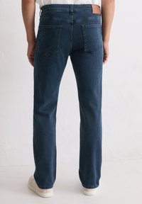 Personne portant un jean droit bleu foncé et des baskets blanches, adossée à un mur blanc uni.