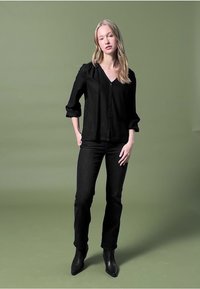 Camicia nera con bottoni e maniche arricciate, abbinata a jeans scuri e stivali neri alla caviglia, su uno sfondo verde smorzato.