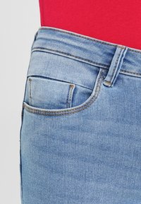 Jeans de mezclilla azul claro con un pequeño bolsillo delantero, una cintura de altura media y costuras en contraste con una parte superior roja sólida.